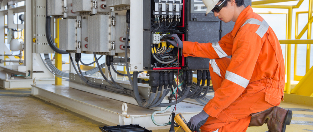 Electrician in orange workwear inspecting electrical panel with multimeter, emphasizing practical support in electrical estimating software.